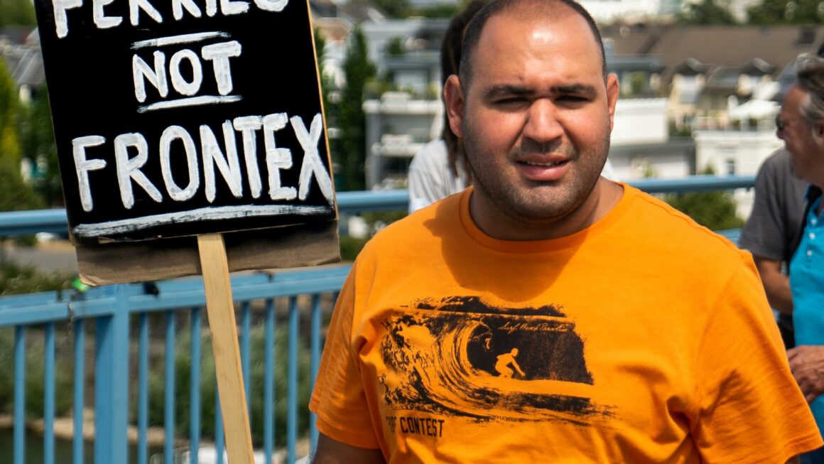 A man with a t-shirt and surfer riding wave image holds a sign stating 'Ferries not Frontex' before a water bay area. He is squinting toward toward the viewer in sunlight.