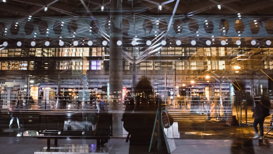 Image of The Hague Centraal train station. A person's reflection in the glass doors of the train station shop lights.