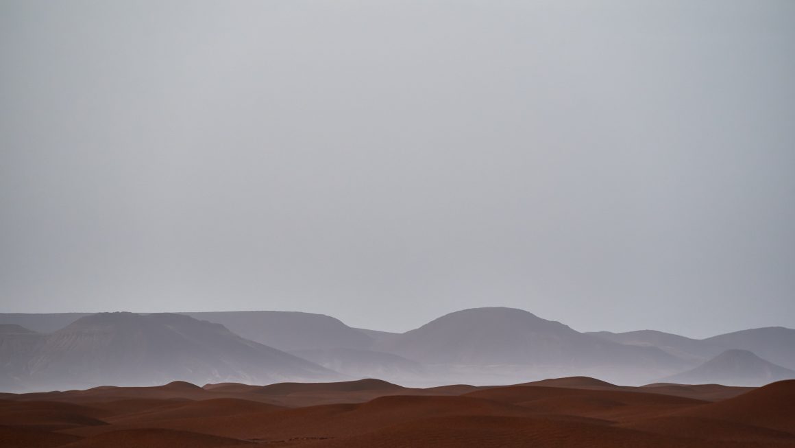 Desert dunes and far mountains shadow under grey foggy sky.