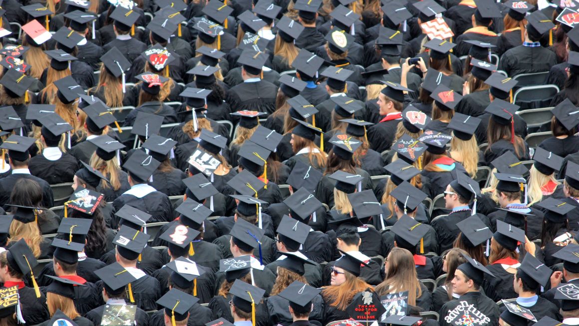 Image of graduates in cap and gowns, sitting in rows on folding chairs at a graduation.