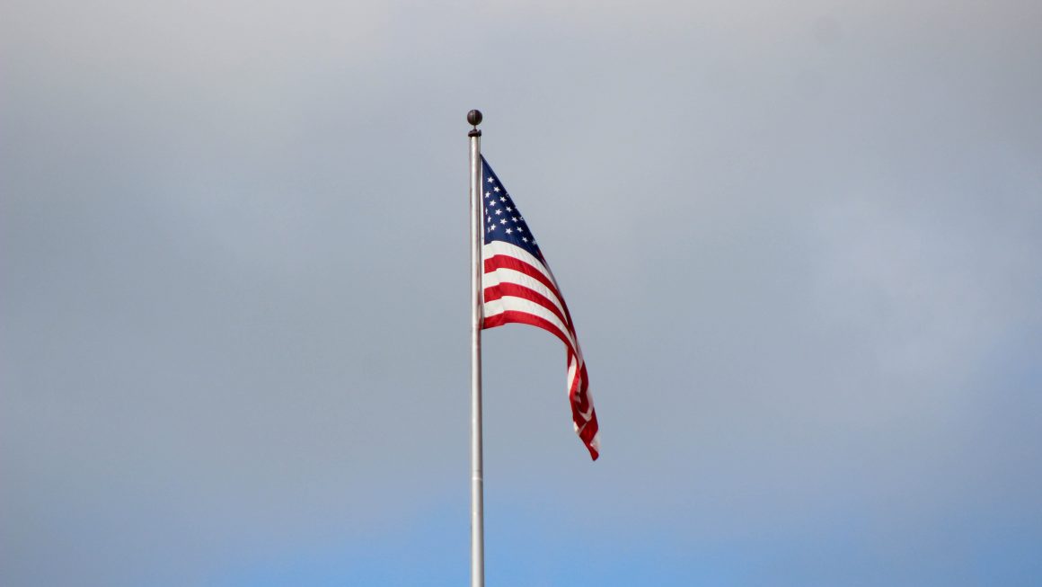 United States' flag on flagpole, in front of sky with clouds.
